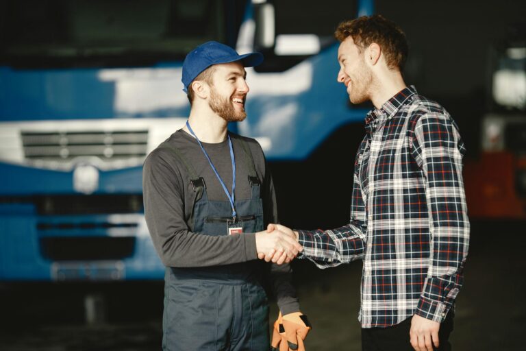 Mechanic in uniform and customer in plaid shirt shaking hands in front of a blue truck inside a garage.